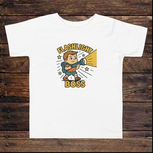 May include: White t-shirt featuring a cartoon illustration of a boy holding a flashlight. The design includes the text "FLASHLIGHT BOSS" in a yellow and black color scheme, with stars and light rays.