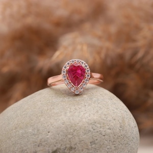 May include: Rose gold ring featuring a pear-shaped, vibrant red gemstone, surrounded by a halo of smaller, clear stones. The ring is set on a smooth, gray stone, with a blurred, neutral background.