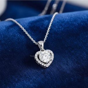Könnte beinhalten: Eine silberne Halskette mit herzförmigem Anhänger. Der Anhänger hat einen großen, runden, klaren Edelstein, der von einem Halo aus kleineren Edelsteinen umgeben ist. Die Halskette ist filigran und silberfarben. Der Hintergrund ist ein dunkelblauer Stoff.