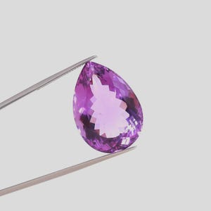 Puede incluir: Una gema de amatista facetada en forma de pera, sujeta con pinzas plateadas. La gema muestra una gama de tonos púrpuras, de violeta claro a oscuro. El corte revela facetas internas, realzando su brillo. Fondo gris neutro.