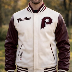 Peut inclure: Veste de baseball Phillies crème avec des manches en cuir marron. La veste présente le logo et le nom de l'équipe Phillies en marron. Elle a un col, des poignets et un ourlet rayés marron et blancs. La veste a deux poches avant et une fermeture boutonnée.