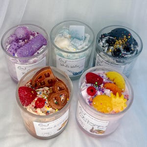 May include: Five dessert-themed candles in clear glass jars. The candles feature various toppings, including waffles, pretzels, and fruit. The labels read "Vanilla & Caramel", "Pink Champagne & Ice Fruit", "White Water", and "Be Calm".