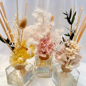 Könnte beinhalten: Drei Glas-Schilf-Diffusoren mit Blumenarrangements. Jede quadratische Flasche hat einen Holzkragen und ist mit Flüssigkeit gefüllt. Die Blumenarrangements umfassen getrocknete Blumen in Gelb, Rosa und Creme, mit Schilfstäbchen.