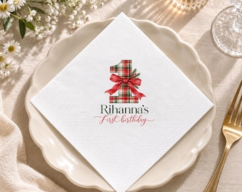 May include: A white paper napkin featuring a large number one with a red and green plaid pattern, adorned with a red ribbon bow and pine sprigs. Below the number, the text reads Rihanna's first birthday in black and red script.