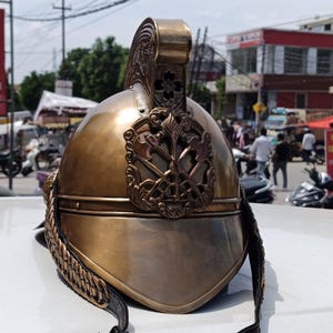 Puede incluir: Un casco de color latón con detalles intrincados, con una cresta y un emblema decorativos. El casco tiene una forma curva y una correa de barbilla de cuero. El diseño sugiere un propósito histórico o ceremonial.