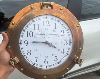May include: A round clock with a brass-colored frame, resembling a ship's porthole. The clock face is white with black numerals and hands. The text "Franklin & Murphy EST. 1868" is printed on the clock face.