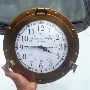Può includere: Un orologio rotondo color ottone, con quadrante bianco e numeri neri. L'orologio ha un design nautico a oblò con la scritta "Franklin & Murphy EST. 1868". L'orologio è tenuto da una persona.