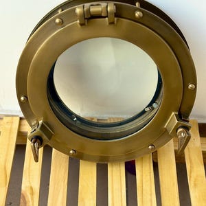 Op de afbeelding: Een messingkleurig, rond patrijspoortvenster met een heldere glazen ruit. De patrijspoort heeft een cirkelvormig frame met zichtbare klinknagels en een scharnierende bovenkant. Twee vergrendelingshendels zijn zichtbaar aan de zijkanten.