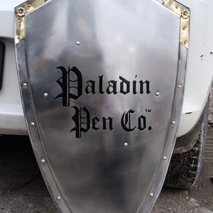 Puede incluir: Un escudo plateado con detalles de latón en las esquinas y remaches. El escudo tiene las palabras "Paladin Pen Co." en una fuente gótica. El escudo está apoyado contra un coche blanco.
