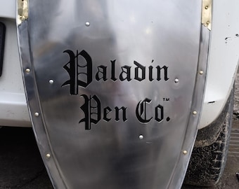 Può includere: Uno scudo color argento con angoli in ottone e rivetti. Lo scudo presenta la scritta "Paladin Pen Co." in un carattere gotico. Lo scudo è appoggiato a un'auto bianca.