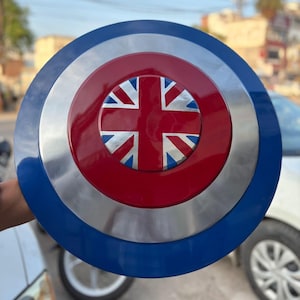 Puede incluir: Un escudo redondo con una combinación de colores azul, plateado y rojo. El centro presenta un círculo rojo con un diseño de la Union Jack. El escudo tiene un acabado brillante y parece ser de metal.