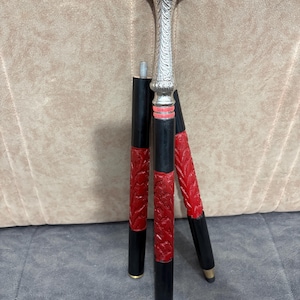 May include: A silver-colored, ornate walking cane with a decorative handle and three black and red legs. The legs have a carved leaf pattern and gold-colored tips. The cane is designed to stand on its own.