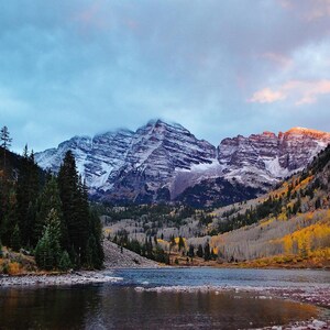 May include: A scenic landscape featuring a lake reflecting the sky and surrounding mountains. The mountains have snow-capped peaks and are flanked by evergreen forests and trees with yellow foliage. The sky is a mix of blue and pink hues.