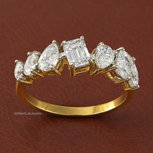 May include: A gold ring featuring a central emerald-cut diamond flanked by two pear-cut diamonds and two smaller round-cut diamonds on each side. The band is polished yellow gold, and the setting includes the text BrilliantLabJewels in small print.