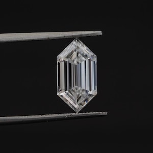 May include: A clear, hexagonal-cut gemstone held by tweezers against a black background. The gem has a symmetrical shape with multiple facets, reflecting light. The gemstone's clarity and cut are prominent.
