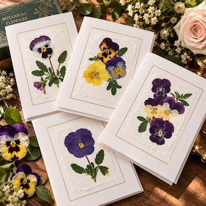 May include: Four white greeting cards featuring pressed pansy flowers with green leaves. Each card has a gold rectangular border and a textured paper inlay. The flowers display purple, yellow, and white petals. The cards are arranged on a surface.