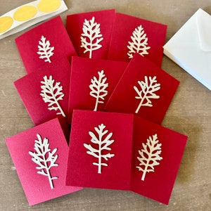 May include: A stack of ten red folded cards with white paper cut-out trees on each card. The cards are arranged in a semi-circle.