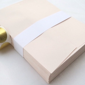 K&ouml;nnte beinhalten: Ein Stapel hellbeige Umschl&auml;ge, die mit einem wei&szlig;en Band zusammengebunden sind. Links ist eine Rolle goldener Folienaufkleber mit schwarzer Schrift zu sehen. Die Umschl&auml;ge sind ordentlich gestapelt, was auf Schreibwaren hindeutet.