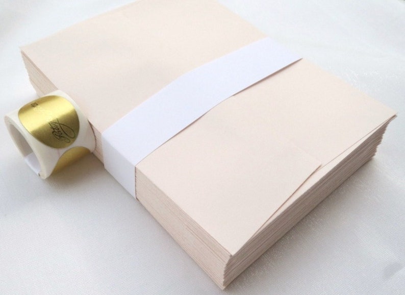 K&ouml;nnte beinhalten: Ein Stapel hellbeige Umschl&auml;ge, die mit einem wei&szlig;en Band zusammengebunden sind. Links ist eine Rolle goldener Folienaufkleber mit schwarzer Schrift zu sehen. Die Umschl&auml;ge sind ordentlich gestapelt, was auf Schreibwaren hindeutet.