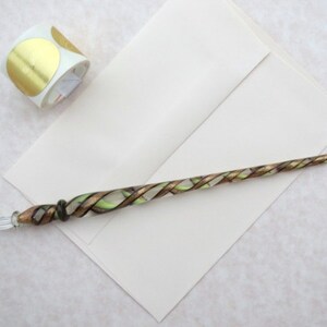 K&ouml;nnte beinhalten: Ein Glas-Dip-Pen mit einem gedrehten Design in Gold, Gr&uuml;n und Klar. Der Stift liegt auf einem wei&szlig;en Umschlag mit einer Rolle goldener Kreisaufkleber im Hintergrund. Der Stift hat eine spitze Feder.