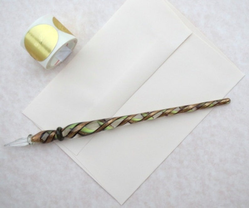 K&ouml;nnte beinhalten: Ein Glas-Dip-Pen mit einem gedrehten Design in Gold, Gr&uuml;n und Klar. Der Stift liegt auf einem wei&szlig;en Umschlag mit einer Rolle goldener Kreisaufkleber im Hintergrund. Der Stift hat eine spitze Feder.