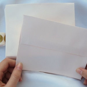 K&ouml;nnte beinhalten: Ein Stapel cremefarbener Umschl&auml;ge mit einem passenden ge&ouml;ffneten Umschlag. Eine Rolle goldener Folienaufkleber ist im Hintergrund zu sehen. Die Umschl&auml;ge bestehen aus strukturiertem Papier.