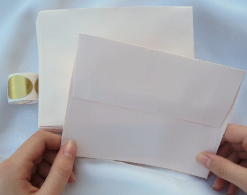 K&ouml;nnte beinhalten: Ein Stapel cremefarbener Umschl&auml;ge mit einem passenden ge&ouml;ffneten Umschlag. Eine Rolle goldener Folienaufkleber ist im Hintergrund zu sehen. Die Umschl&auml;ge bestehen aus strukturiertem Papier.
