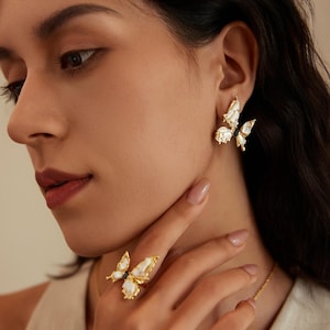 May include: Gold-toned butterfly earrings and ring set. The jewelry features white and black accents, with a detailed butterfly design. The ring is on a finger, and the earrings are on an ear. The jewelry is set against a soft background.