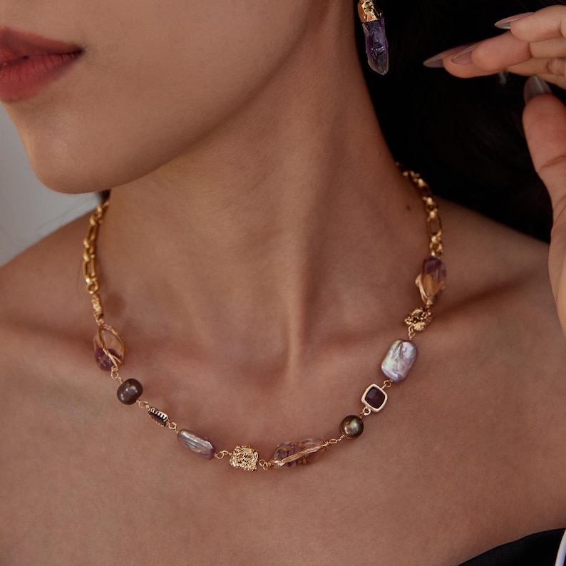 May include: A gold-toned necklace with a variety of gemstones, including purple amethyst, pearls, and dark brown beads. The necklace has a chain link design and is paired with matching earrings. The jewelry is displayed on a person.