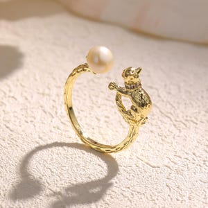May include: A gold-colored ring featuring a cat design. The ring has a pearl at the top and a cat figure with a purple gem collar. The ring is set against a textured, off-white surface.
