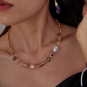 May include: A gold-toned necklace with a variety of gemstones and pearls. The necklace features a mix of purple, brown, and clear stones, along with a few pearls. A matching earring with a purple stone hangs nearby.