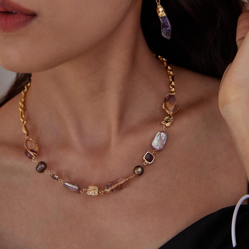 May include: A gold-toned necklace with a variety of gemstones and pearls. The necklace features a mix of purple, brown, and clear stones, along with a few pearls. A matching earring with a purple stone hangs nearby.