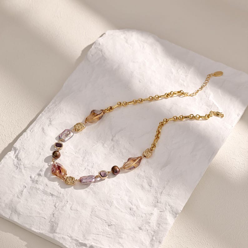 May include: A gold-toned necklace with a variety of beads in shades of brown, purple, and clear. The necklace features a delicate chain and decorative gold accents. The beads have an irregular shape, adding to the necklace's unique design.