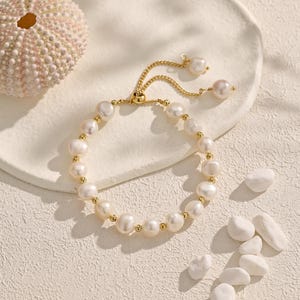 May include: A gold-toned bracelet featuring a series of white pearls and small gold beads. The adjustable bracelet has a delicate chain and is displayed on a white surface with decorative elements.
