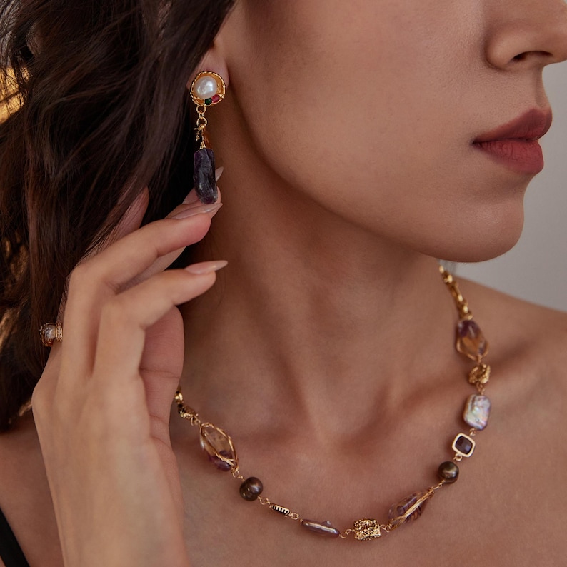 May include: Gold-toned jewelry set featuring a necklace and earrings. The necklace has a chain with various shaped stones in shades of purple, brown, and clear. The earrings have a pearl, red and green stones, and a dark purple stone.