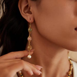 May include: Gold-toned drop earrings featuring three encased, colorful gemstones and a dangling pearl. The gemstones are in shades of green, pink, and yellow. The earrings are suspended from a delicate gold wire.