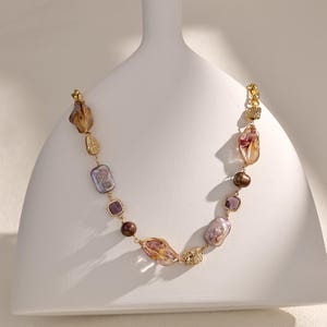 May include: A gold-toned necklace featuring a variety of irregularly shaped, colorful beads. The necklace includes large, translucent amber-colored stones, iridescent purple-gray rectangles, and round brown beads. Accents include textured gold-colored elements.