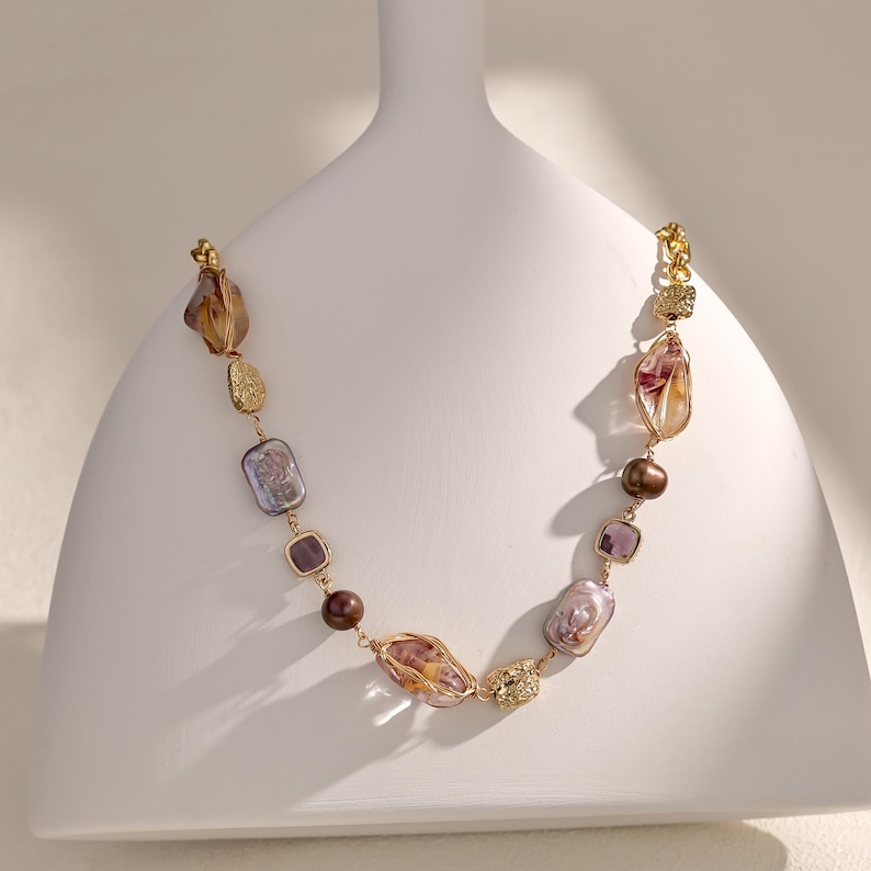 May include: A gold-toned necklace featuring a variety of irregularly shaped, colorful beads. The necklace includes large, translucent amber-colored stones, iridescent purple-gray rectangles, and round brown beads. Accents include textured gold-colored elements.