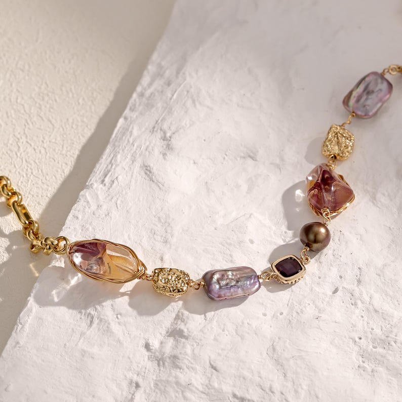 May include: A gold-toned necklace featuring a variety of irregularly shaped, colorful beads. The beads include shades of purple, amber, and gold, with a mix of textures. The necklace has a delicate chain and clasp.