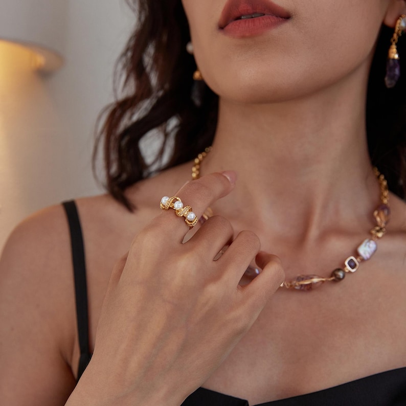 May include: Gold-toned ring featuring three white pearls and delicate detailing. The ring is worn on a finger, showcasing its elegant design. A matching necklace with pearls and gemstones is also visible.