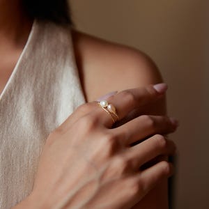 May include: A delicate gold ring featuring two freshwater pearls, one white and one pink. The ring's design includes a twisted band, adding a touch of texture and elegance. The pearls are the focal point, creating a simple yet sophisticated piece of jewelry.