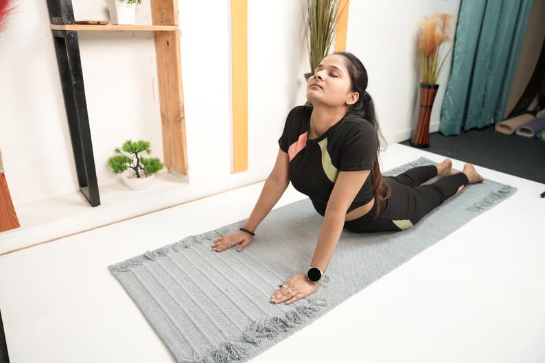 May include: A person in a black athletic outfit is doing yoga on a grey yoga mat with fringe. The outfit has pink and green accents. A small bonsai tree and decorative shelf are in the background.