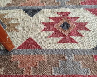 May include: Close-up of a woven rug with a geometric pattern. The rug features a mix of colors including red, beige, gray, and brown. The design includes diamond and triangle shapes, creating a textured, rustic look.