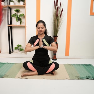 May include: A person in a black yoga outfit with pink and green accents sits in a meditative pose on a striped yoga mat. The mat transitions from green to beige. Decorative vases and plants are in the background.