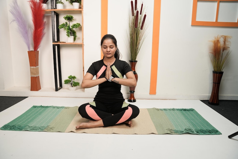 May include: A person in a black yoga outfit with pink and green accents sits in a meditative pose on a striped yoga mat. The mat transitions from green to beige. Decorative vases and plants are in the background.