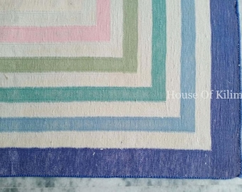 Può includere: Un tappeto rettangolare con un motivo geometrico. Il tappeto presenta un bordo blu scuro, con strisce alternate bianche, rosa, verdi e azzurre. Il testo "House Of Kilims" è visibile nell'angolo.