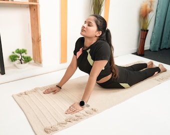 May include: A woman in a yoga pose on a beige rectangular yoga mat. She is wearing a black athletic outfit with pink and green accents. The mat has a textured surface and tassels on the ends. A small bonsai tree is visible in the background.