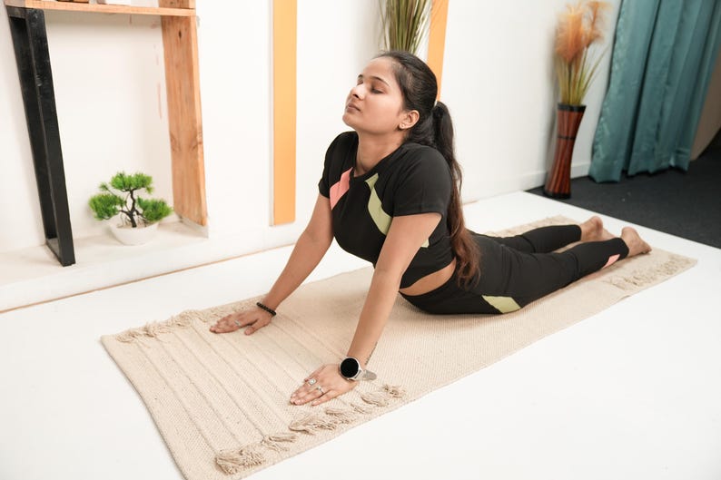 May include: A woman in a yoga pose on a beige rectangular mat. She wears a black athletic outfit with pink and green accents. Her arms are extended, and her eyes are closed. A small bonsai tree and a wooden shelf are in the background.