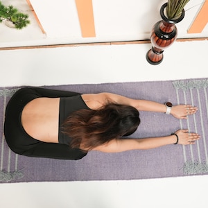 May include: A person in a yoga pose on a lavender and white striped yoga mat with fringe. The person is wearing a black sports bra and black leggings. A watch and bracelet are visible.
