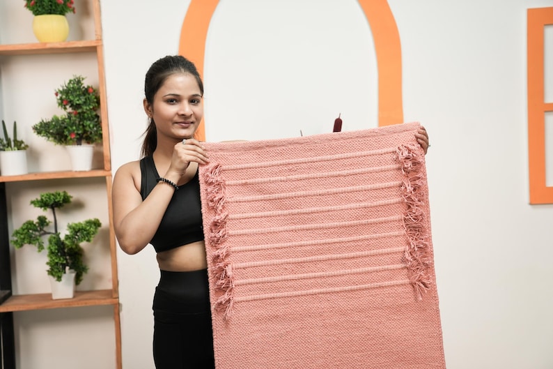 May include: A rectangular, peach-coloured rug with horizontal ribbed detailing and fringed edges. The rug is held up, showcasing its texture and design. The rug is likely used for home decor or as a floor covering.